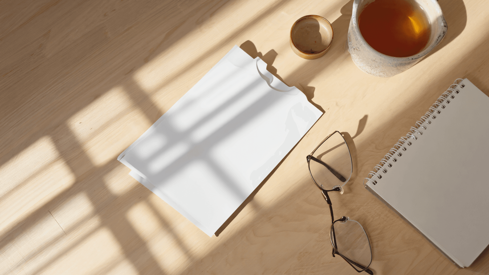 A printed leaflet, notebook, reading glasses and a mug of tea on a sunlit desk